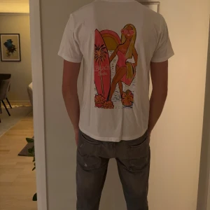 Vit t-shirt med Beach Babe-tryck - Säljer en vit t-shirt i bomull med färgglatt Beach Babe-tryck på ryggen. Trycket har motiv av en tjej med solglasögon, surfbräda och blommor i rosa, orange och gul. Klassisk rund hals och korta ärmar. Perfekt för dig som gillar sommarvibbar och coola prints.