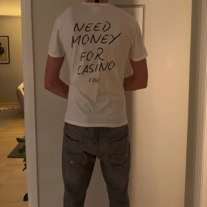 Vit t-shirt Gildan med tryck - Vit t-shirt från Gildan Softstyle med svart handskrivet tryck på ryggen: 'Need money for casino'. Rund hals och klassisk passform. Materialet är mjuk bomull och märkt med Atire. Perfekt för dig som gillar statement-plagg.