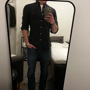 Svart skjorta från Ralph Lauren M - Svart skjorta från Ralph Lauren i slim fit med klassisk krage och vita knappar. Broderad röd logga på bröstet ger en snygg kontrast. Skjortan är långärmad och tillverkad i mjuk bomull, perfekt för dig som gillar stilrena och tidlösa plagg.