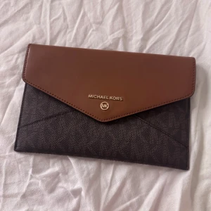 Michael Kors brun clutch - Snygg kuvertväska från Michael Kors i brunt och mörkbrunt med logomönster. Väskan har ett stilrent kuvertlock i ljusbrunt skinn och guldiga detaljer. Perfekt storlek för det viktigaste och stängs med tryckknapp. Klassisk och trendig design.