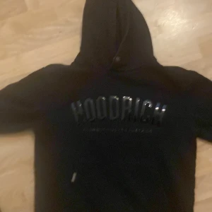 Svart hoodie från Hoodrich - Svart hoodie från Hoodrich med stor, glansig logga på bröstet och texten 'From nothing to something'. Tröjan har huva med knappdetalj och en stor magficka framtill. Perfekt för dig som gillar streetwear och vill ha en clean look.