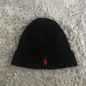 Svart mössa från Polo Ralph Lauren - Svart ribbstickad mössa från Polo Ralph Lauren med röd broderad logga framtill. Klassisk och enkel design som passar perfekt för kalla dagar. Mjuk och skön kvalitet med uppvikt kant.