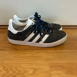 Adidas Gazelle sneakers mörkblå - Säljer ett par klassiska Adidas Gazelle sneakers i mörkblå mocka med vita detaljer och snörning. Skorna har de ikoniska tre ränderna på sidan, guldtexten 'GAZELLE' och vit platt sula. Adidas-logga på hälen och tungan. 