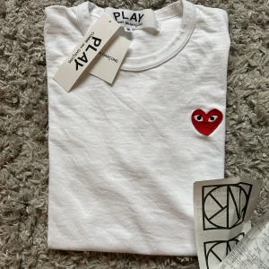 Vit Comme des Garçons Play t-shirt - Vit t-shirt från Comme des Garçons Play med det ikoniska röda hjärtat med ögon broderat på bröstet. Klassisk rund hals och korta ärmar. Tillverkad i mjuk bomull för en skön känsla. Perfekt för dig som gillar stilrena och trendiga plagg. Köpte på NK och använde en gång men när jag använde den kändes den lite liten så säljer den därför. Skulle säga att den är lite liten i storlek och mer som S