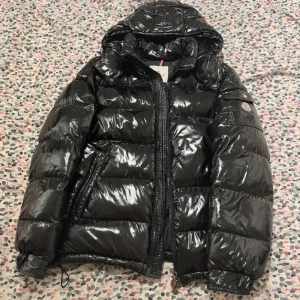 Svart Moncler Maya  - Säljer en svart, glansig pufferjacka från Moncler med huva och dragkedja. Jackan har klassisk Moncler-logga på ärmen, stora fickor och quiltad design. Perfekt för kalla dagar när du vill hålla stilen och värmen.