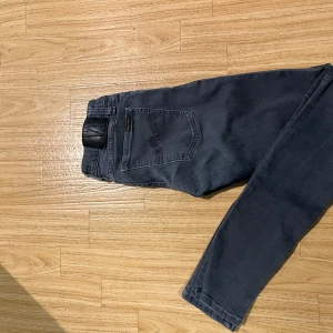 Mörkgrå Nudie Jeans  28/32 - Snygga mörkgrå jeans från Nudie Jeans i skinny fit. Klassisk femficksmodell med svarta detaljer och Nudie-logga på bakfickan. Jeansen är tillverkade i stretchig bomull för skön passform och har raka ben med smal siluett.