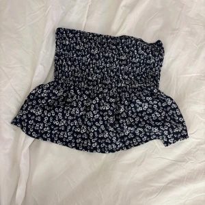 Blommig mörkblå bandeau tubtopp - Supersöt bandeau tubtopp i mörkblått med små vita blommor. Toppen har smockad överdel som sitter tight och en lätt volang nedtill för extra flow. Man kan använda plagget till både topp och kjol. Använd 1 gång! Från Gina🌷