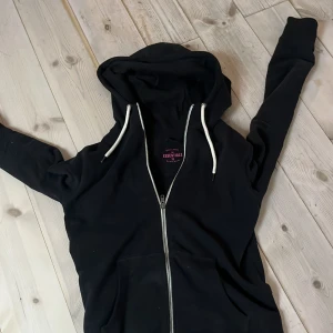 Svart hoodie från Race Marine Essentials - Svart hoodie med dragkedja och vita snören från Race Marine Essentials. Tillverkad i ekologisk bomull, har huva och praktisk känguruficka framtill. Perfekt för chill dagar och streetstyle. Långärmad och med normal passform.