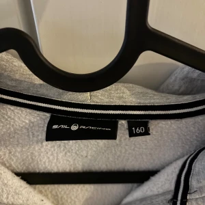 Grå hoodie från Sail Racing med dragkedja - Snygg grå hoodie från Sail Racing med hel dragkedja och logga på bröstet. Tröjan har huva med broderad detalj, ribbad kant vid halsen och långärmade ärmar. Perfekt för chill dagar och har en sportig vibe.