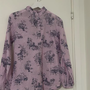 Ljuslila blommig skjorta med volangkrage - Säljer en ljuslila skjorta med mörkblå blommigt mönster och diskreta ränder. Skjortan har en söt volangkrage, knappar framtill och långa ärmar. Materialet känns lätt och luftigt, perfekt för dig som gillar romantisk stil och detaljer.