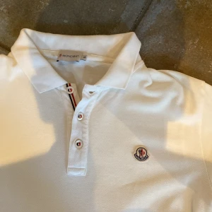 Vit Moncler pikétröja - Vit pikétröja från Moncler med klassisk krage, knappar framtill och Moncler-logga på bröstet. Tricolore-detalj vid knappslån ger en snygg touch. Tillverkad i mjuk bomull och har korta ärmar för en clean och stilren look.