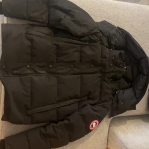 Svart dunjacka från Canada Goose - Säljer en svart dunjacka från Canada Goose med klassisk patch på ärmen. Jackan har huva, dragkedja och knappar framtill samt stora fickor. Perfekt för kalla dagar och riktigt snygg streetstil.