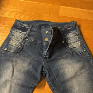 Blå skinny jeans med nitar från Versus - Snygga blå jeans med skinny passform från Versus, dekorerade med silvriga nitar på både fram- och bakfickor. Hög midja och klassisk femficksdesign. Jeansen har slitningar och tvättade detaljer för en cool look. Saknas en knapp fram