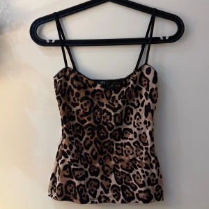 Leopardmönstrad linnetopp från Zara - Säljer en snygg leopardmönstrad linnetopp från Zara i beige och svart med tunna axelband. Toppen har en liten svart rosett framtill och dekorativ spetskant längs halsringningen. Materialet är glansigt och känns lyxigt, perfekt för att sticka ut.