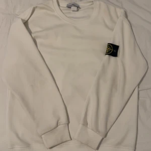 Vit sweatshirt från Stone Island - Vit sweatshirt från Stone Island med klassisk rund hals och ribbade muddar. Tröjan har den ikoniska svarta logotyp-patchen på vänster ärm. Tillverkad i mjuk bomull, perfekt för en clean och stilren look.