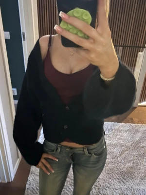 Svart cropped kofta med knappar - Trendig svart cropped kofta med knappar framtill och långa, lite pösiga ärmar. Perfekt att bära över ett linne eller topp för en avslappnad men snygg look. Koftan har en mjuk och fluffig känsla och passar till både jeans och kjol.