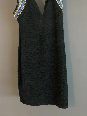 Svart klänning med pärldetaljer - Snygg svart klänning med v-ringning och pärlbroderade axelband i blått, guld och vitt. Klänningen har en rak passform och är ärmlös, vilket ger en stilren och modern look. Perfekt för dig som vill sticka ut med detaljerade axlar.