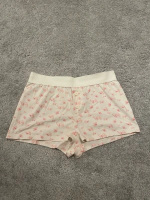 Pyjamas shorts - Säljer ett par krämfärgade loungewear-shorts med rosa fjärilsmönster och bred resår i midjan. Framtill finns tre dekorativa knappar. Perfekta för mysiga hemmakvällar och avslappnade dagar. Bara testade och inga skador, köpa för original pris.