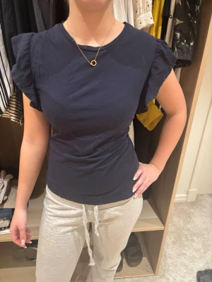 Mörkblå topp med volangärm Gina Tricot - Mörkblå topp från Gina Tricot med volangärmar och rund halsringning. Tillverkad i mjuk bomull som känns skön mot huden. Passformen är normal och toppen har en enkel, stilren look som passar till många outfits.