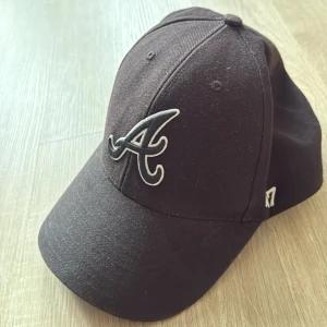 Svart keps med Atlanta Braves-logga - Snygg svart keps med böjd skärm och broderad vit Atlanta Braves-logga på framsidan. Justerbar snapback baktill för perfekt passform. Tillverkad i bomullsmaterial och har klassisk basebollkeps-design.