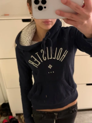 Fodrad Hollister hoodie  - Mysig mörkblå hoodie från Hollister med vit broderad logga på bröstet. Hoodien har en fodrad huva med fluffigt vitt material och dragsko, samt en klassisk känguruficka framtill. Perfekt för en chill och avslappnad stil.