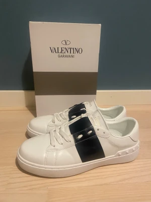 Valentino Garavani vita sneakers - Snygga vita sneakers från Valentino Garavani med bred mörkblå rand och nitar på hälen. Skorna har snörning och en klassisk låg siluett. Tillverkade i skinn för en lyxig känsla och stilren look.