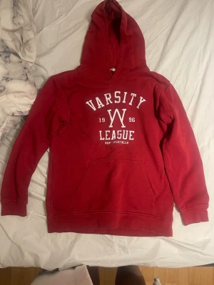 Röd hoodie Varsity League - Röd hoodie från Varsity League med stor vit logga och text framtill. Klassisk känguruficka och mjukt material på insidan. Huva utan dragsko och ribbade muddar vid ärmar och nederkant. Perfekt för en avslappnad och sportig stil.  Tveka inte att höra av dig om du har några frågor, snabb leverans 📦 💕