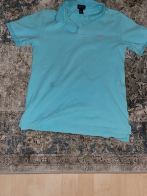 Turkos pikétröja från Polo Ralph Lauren - Snygg turkos pikétröja från Polo Ralph Lauren med klassisk krage och knappslå. Tröjan har den ikoniska rosa logotypen broderad på bröstet och är tillverkad i mjuk bomull. Perfekt för en avslappnad och stilren look.