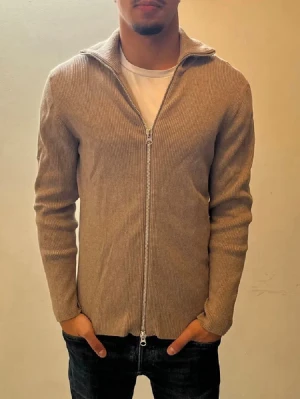 Beige ziptröja från Ljung by Marcus Larsson - Säljer en beige ribbad ziptröja från LJUNG by Marcus Larsson. Tröjan har hel dragkedja, hög krage och är tillverkad i ett mjukt stickat material. Perfekt för lager på lager och enkel att matcha med olika outfits.