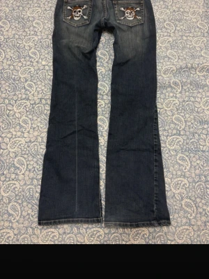 Mörkblå bootcut jeans med döskalle - Säljer ett par mörkblå bootcut jeans med slitningar och broderade döskallar på bakfickorna. Jeansen har klassisk femficksmodell och coola detaljer som ger en edgy vibe. Perfekta för dig som vill sticka ut lite extra. Storlek 42 men passar även 40 och 38