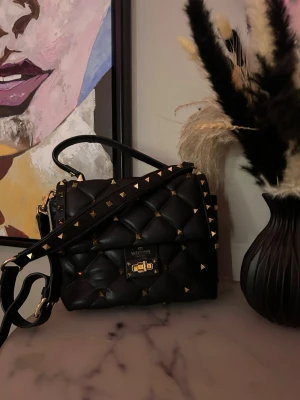 Svart quiltad handväska Valentino - Har bestämt mig för att sälja min snygga svart quiltad handväska från Valentino med guldfärgade nitar och detaljer. Väskan har både handtag och axelrem, samt ett spänne framtill. (Går att ta av axelbandet). Tillverkad i skinn och har en lyxig känsla med coola detaljer. Helt äckta men vintage modell. Går att vrida på nitarna! As snygg design, perfekt för dig som vill ha en unik accessoar med dig. Väskan har en slitning vid låset men inte märkbart. Annars är väskan väldigt försiktigt använd!❤️ 
