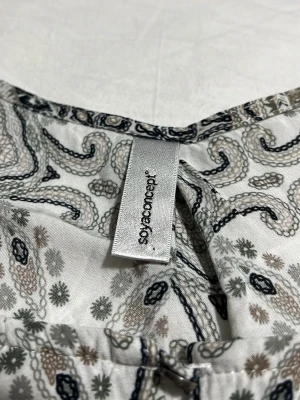 Mönstrad ärmlös topp Soyaconcept - Snygg ärmlös topp från Soyaconcept med detaljerat grafiskt mönster i svart, grått och beige på vit botten. Lätt och luftig modell med rund halsringning, perfekt för en trendig och avslappnad look.