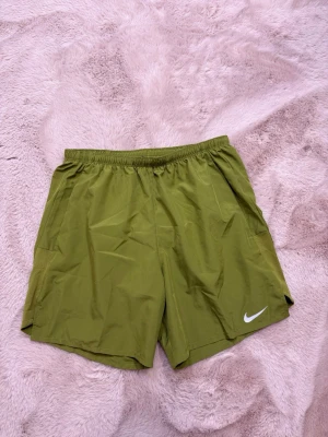 Nike - Snygga gröna träningsshorts från Nike med elastisk midja och vit logga på benet. Tillverkade i lätt och snabbtorkande syntetmaterial, perfekta för aktiva dagar. Klassisk passform och enkel design som funkar till det mesta.