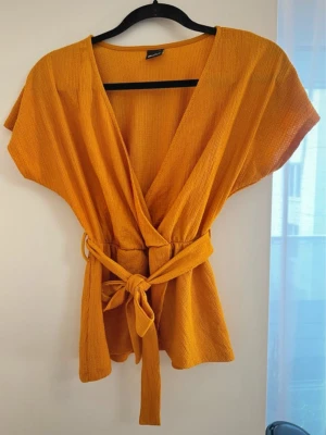 Blus från Ginatricot - Snygg gul omlottopp med korta ärmar och breda knytband i midjan. Toppen har v-ringad hals och ett lätt strukturerat tyg som ger en avslappnad men trendig look. Perfekt att styla med jeans eller kjol för en cool vibe. Passar även S.