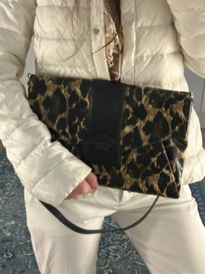Leopardmönstrad kuvertväska från Victoria’s secret - Snygg kuvertväska från Victoria’s secret i leopardmönstrat skinn med svarta detaljer. Väskan har en stilren rektangulär form och ett brett svart band över locket samt en praktisk handledsrem. Perfekt accessoar för att lyfta din outfit.