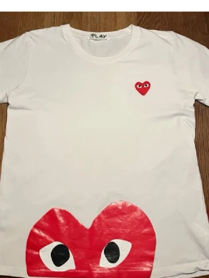 Vit Comme des Garçons Play t-shirt - Vit t-shirt från Comme des Garçons Play med det ikoniska röda hjärtat med ögon på bröstet och ett stort hjärtmotiv längst ner. Klassisk passform och rund halsringning. Perfekt för dig som gillar streetwear och vill sticka ut.