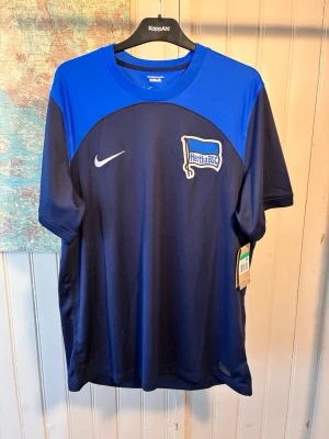 Hertha B.S.C. Nike 2022/23 away kit, size XL - Brand new  #lukebakio #piatek #herthabsc #bundesliga #germany #nike  Här är en fotbollströja från Nike med Hertha BSC-emblem på bröstet. Tröjan har en blå färg på axlarna och en mörkare blå färg på resten av tröjan. Den har även en vit Nike-logga på bröstet.