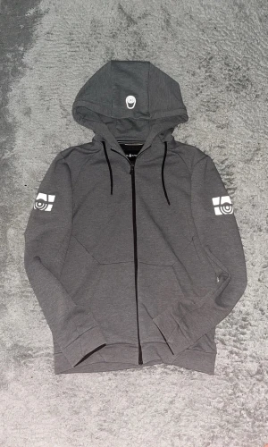 Grå hoodie med tryck på ärmarna - Snygg grå hoodie med dragkedja och svart snörning i huvan. Har vita tryck på båda ärmarna och en liten vit detalj på huvan. Perfekt för en avslappnad streetstyle-look.