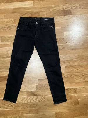 Svarta Replay Anbass jeans - Säljer ett par svarta Replay Anbass jeans med klassisk femficksdesign och smal passform. Jeansen har en snygg svart tvätt, diskret Replay-logga på bakfickan och patch på linningen. Perfekta för dig som gillar stilrena och moderna jeans.