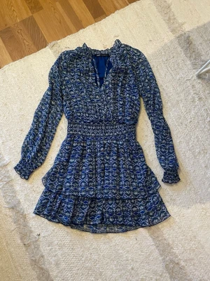 Blå mönstrad kortklänning med volang - Supersöt blå kortklänning med vitt mönster och volangdetaljer. Klänningen har långärmade ärmar med smock vid handlederna, markerad midja och v-ringad hals. Perfekt för dig som gillar en feminin och trendig stil.