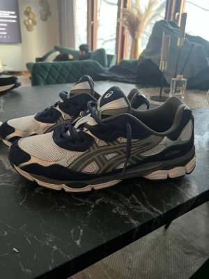 Asics gel nyc - Snygga Asics sneakers i blått, vitt och silver med klassisk mesh och syntetmaterial. Skorna har rund tå, platt sula och tydliga Asics-loggor på sidorna. Perfekta för dig som gillar sportig och trendig stil.