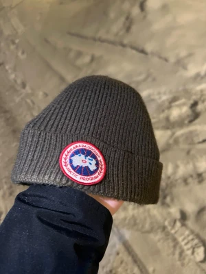 Mörkgrön ribbstickad mössa Canada Goose - Snygg mörkgrön ribbstickad mössa från Canada Goose med klassisk röd och vit logopatch framtill. Tillverkad i 100% bomull och har uppvikt kant för extra stilpoäng. Perfekt för dig som vill ha både värme och streetstyle.