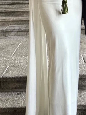 Vit långklänning med oneshoulder - Elegant vit långklänning med figurnära passform och oneshoulder-design. Klänningen har en lång ärm på ena sidan och en snygg cape-detalj som ger en dramatisk look. Perfekt för speciella tillfällen när du vill sticka ut.