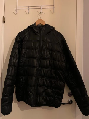 Svart pufferjacka från Funk - Svart pufferjacka från Funk med huva och dragkedja framtill. Jackan har quiltad design och är tillverkad i syntetmaterial. Perfekt för kyliga dagar och enkel att matcha med olika outfits.