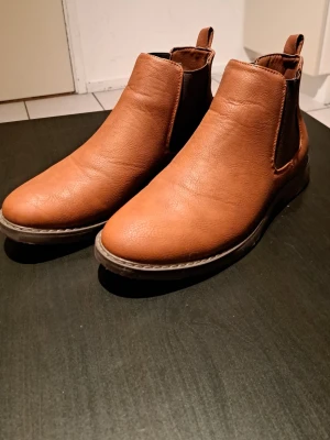 Bruna Chelsea boots från YOURTURN - Snygga bruna Chelsea boots från YOURTURN i skinnimitation med elastiska paneler på sidorna. Klassisk rund tå och låg klack. Perfekta för dig som vill ha stilrena boots med enkel design och bekväm passform.