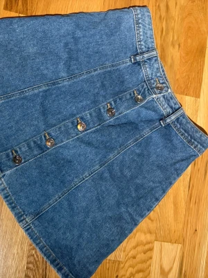 Blå jeanskjol med knappar - Säljer en klassisk blå jeanskjol i kort modell med raka linjer och snygga silverfärgade knappar framtill. Kjolen har hällor för skärp och är gjord i 100% bomull, perfekt för en avslappnad och trendig look.