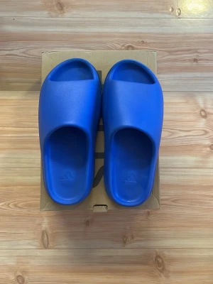 Blå Adidas tofflor Yeezy Slide - Säljer ett par blå Adidas Yeezy Slide med öppen tå och platt sula. Tofflorna är tillverkade i syntetmaterial och har en clean, minimalistisk design med Adidas-logga på fotbädden. Perfekta för chill dagar hemma eller på stranden.