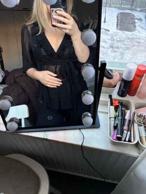 Svart transparent blus med knyt - Säljer en svart transparent blus med v-ringning och knyt i midjan. Blusen har trekvartsärm och volangdetaljer vid ärmslut och längs kanten. Perfekt för dig som gillar en luftig och trendig look.