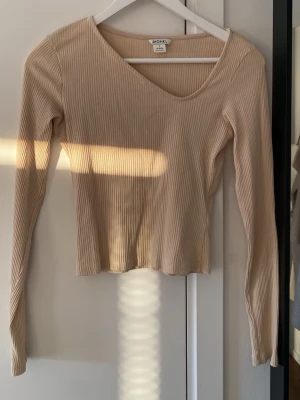 Beige ribbad topp från Monki - Säljer en beige ribbad långärmad topp från Monki. Toppen har en djup v-ringning och är croppad i modellen, storlek S. Använd endast 1 gång!