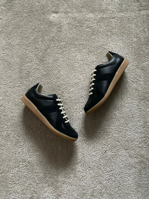 Maison Margiela Gats med NFC - Stilrena svarta Gats sneakers med beige gummisula och vita snören. Klassisk låg modell med rund tå och diskreta paneler i skinn och mocka. Perfekta för dig som gillar minimalistisk och tidlös design. Storlek 44, 29cm innersula